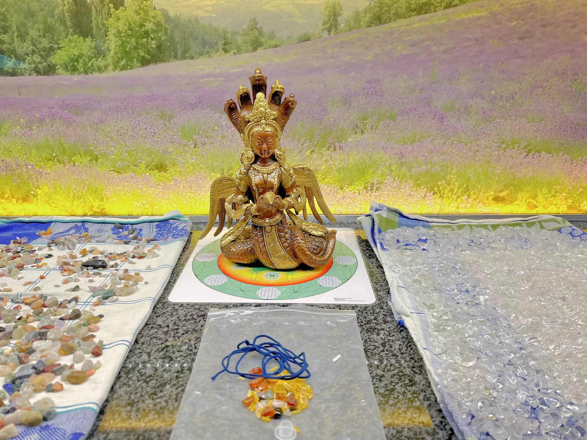 Goldene mehrköpfige Naga‑Statue auf einem farbigen Tuch, flankiert von sortierten Edelsteinen und Kristallen auf zwei Stoffunterlagen, im Hintergrund ein Landschaftsmotiv mit Lavendelfeld.