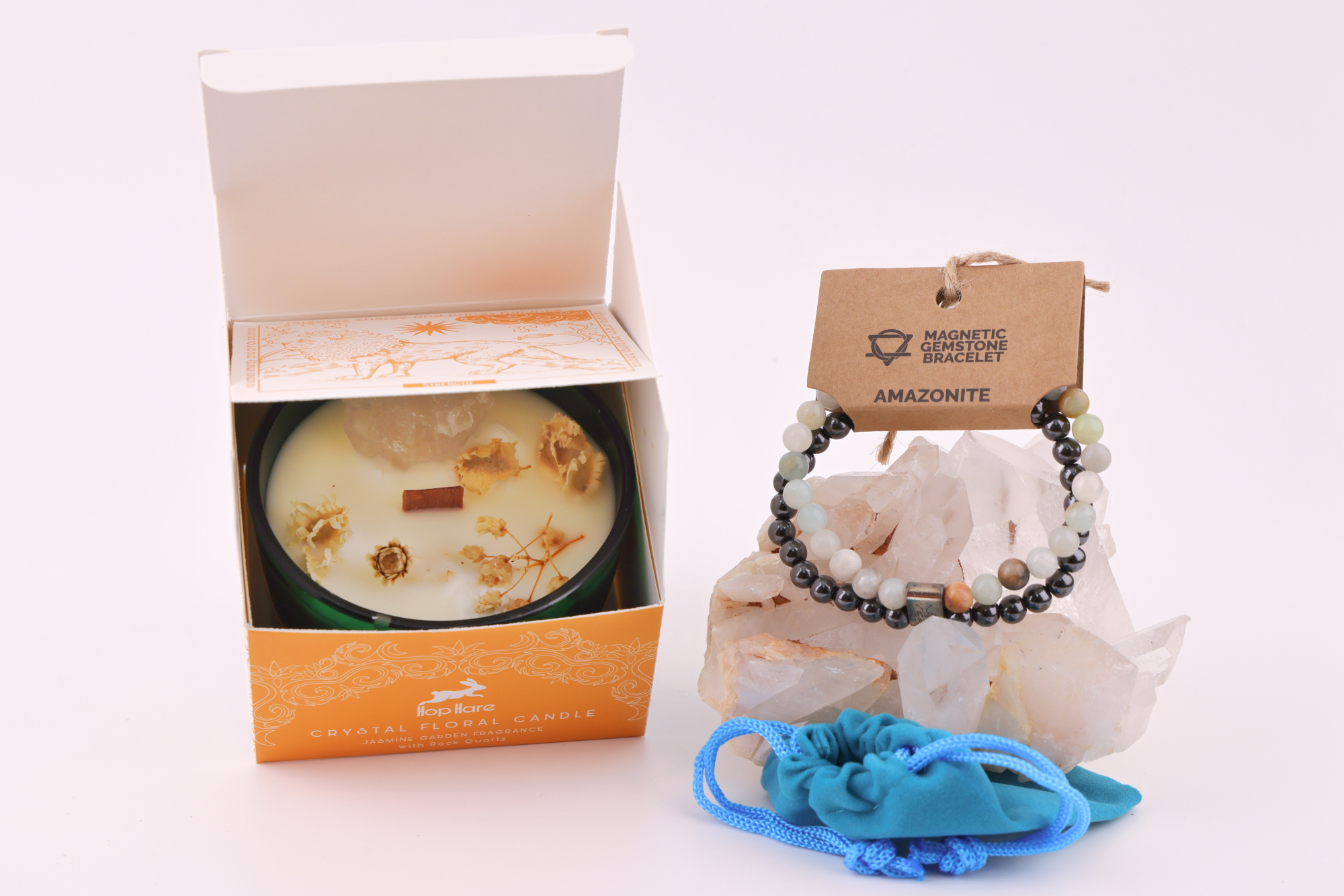 Amazonit‑Magnetarmband auf einem klaren Bergkristall‑Cluster, daneben eine Duftkerze mit floralen Elementen in einer orangefarbenen Verpackung sowie ein blaues Stoffbeutelchen.