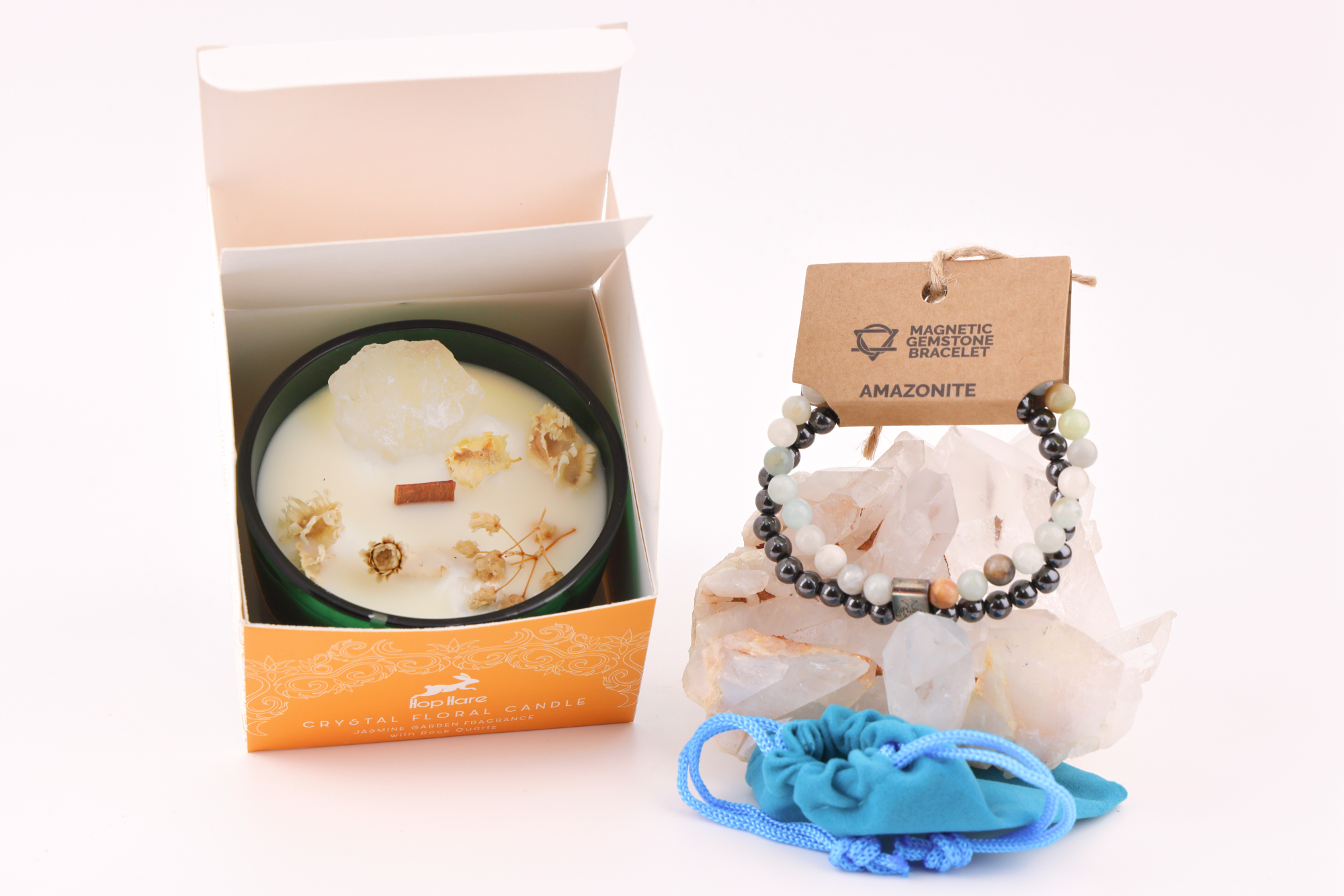 Amazonit‑Magnetarmband auf einem klaren Bergkristall‑Cluster, daneben eine Duftkerze mit floralen Elementen in einer orangefarbenen Verpackung sowie ein blaues Stoffbeutelchen.