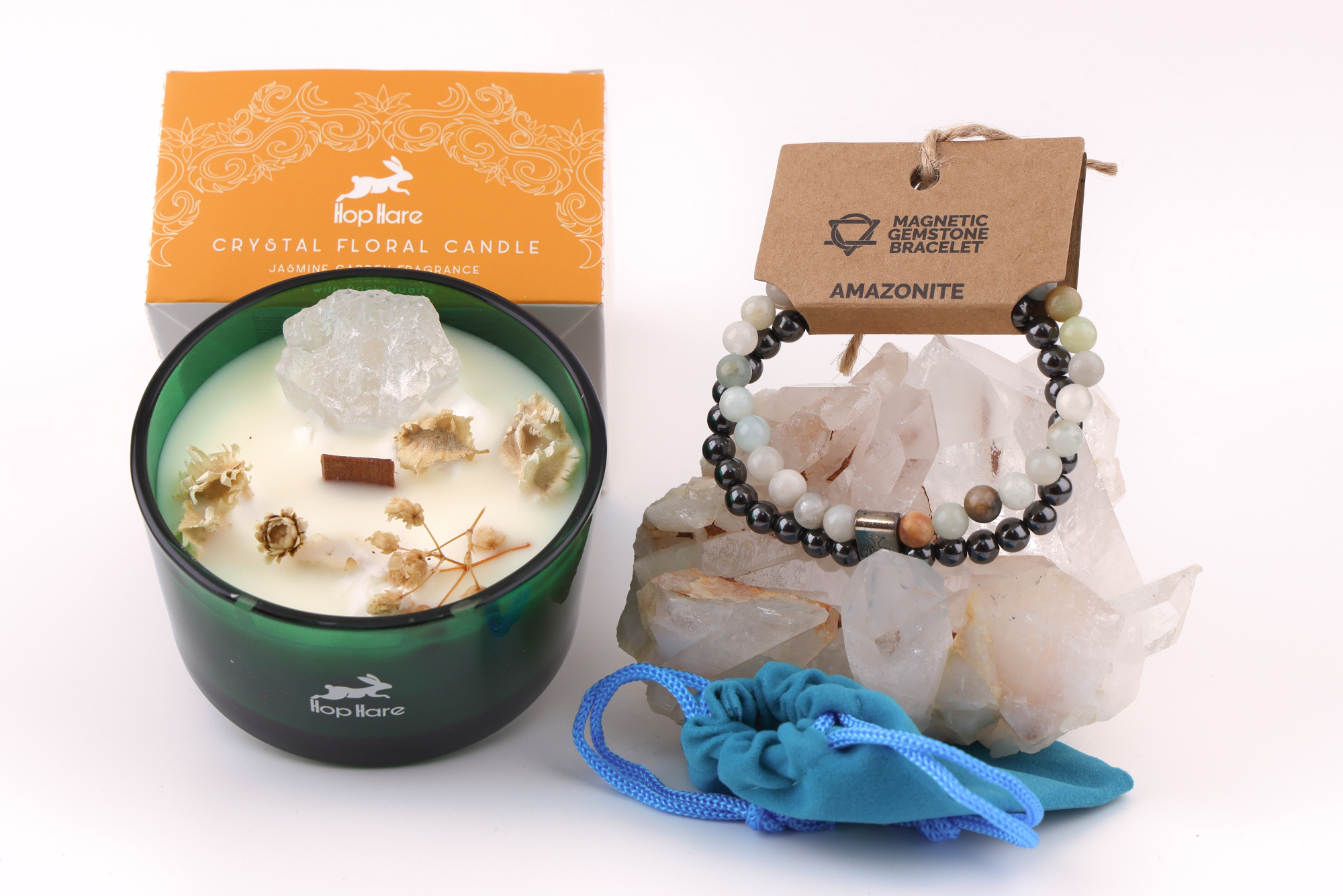 Amazonit‑Magnetarmband auf einem klaren Bergkristall‑Cluster, daneben eine Duftkerze mit floralen Elementen in einer orangefarbenen Verpackung sowie ein blaues Stoffbeutelchen.