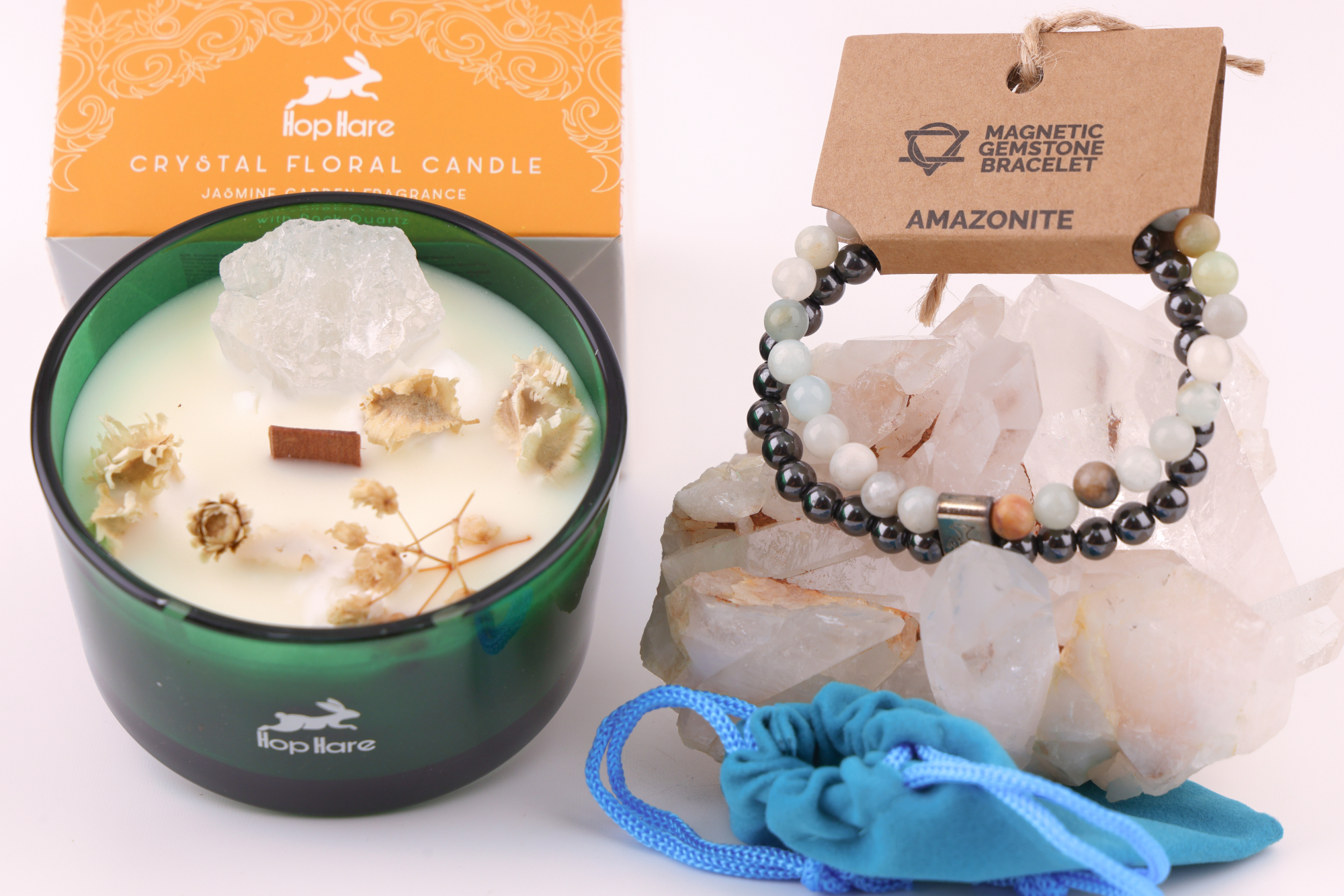 Amazonit‑Magnetarmband auf einem klaren Bergkristall‑Cluster, daneben eine Duftkerze mit floralen Elementen in einer orangefarbenen Verpackung sowie ein blaues Stoffbeutelchen.
