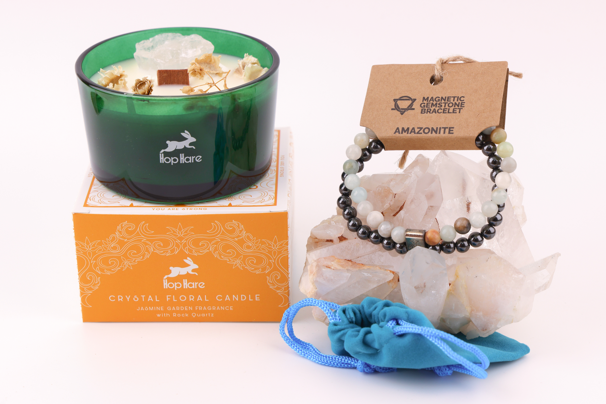Amazonit‑Magnetarmband auf einem klaren Bergkristall‑Cluster, daneben eine Duftkerze mit floralen Elementen in einer orangefarbenen Verpackung sowie ein blaues Stoffbeutelchen.