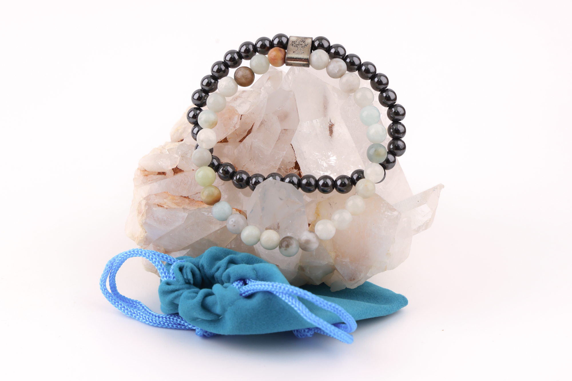 Sodalith‑Magnetarmband mit schwarzen und hellen Edelsteinperlen, dekorativ präsentiert auf einem klaren Bergkristall‑Cluster; davor ein türkisfarbenes Stoffbeutelchen mit blauen Zugbändern.