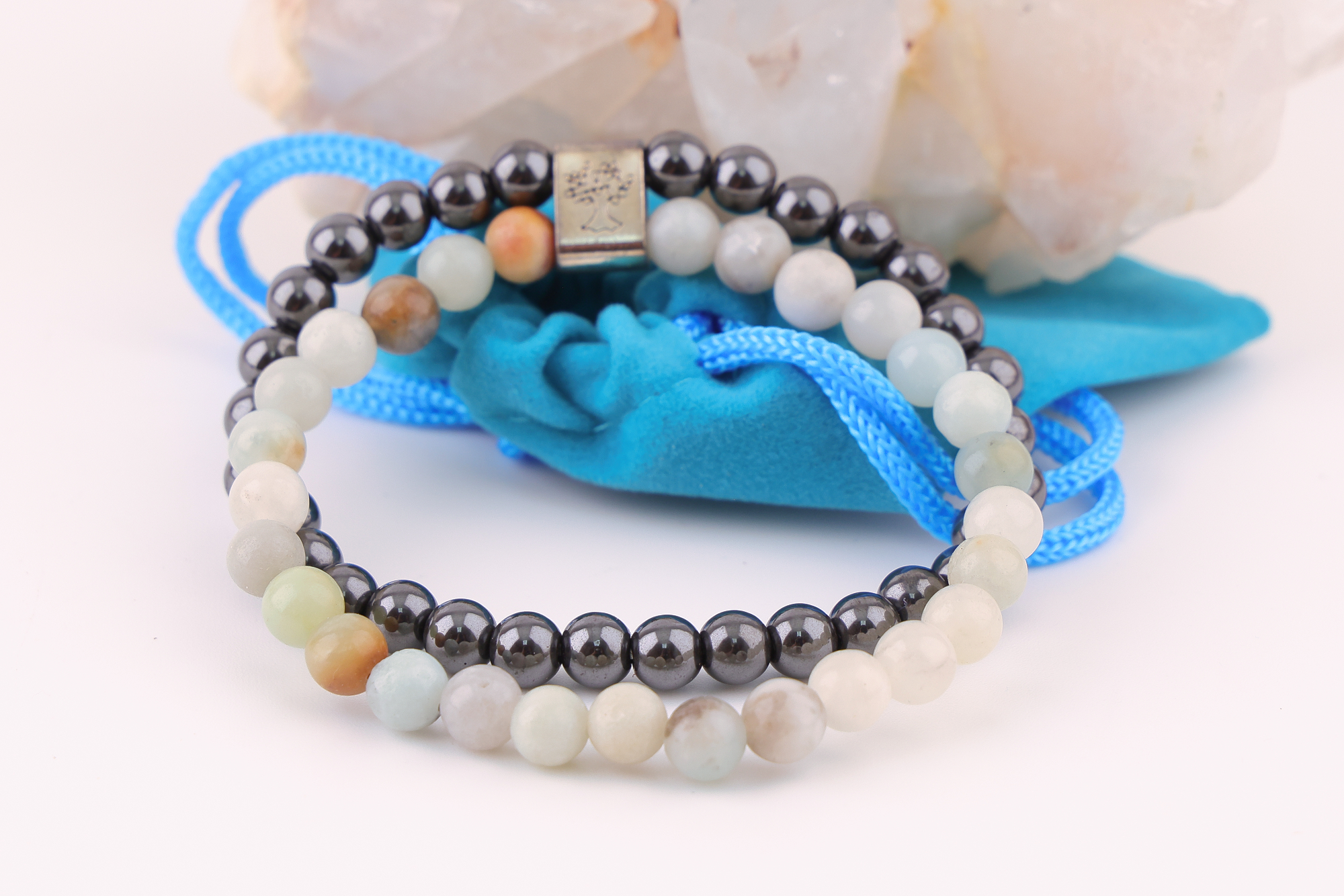 Sodalith‑Magnetarmband mit schwarzen und hellen Edelsteinperlen, dekorativ präsentiert auf einem klaren Bergkristall‑Cluster; davor ein türkisfarbenes Stoffbeutelchen mit blauen Zugbändern.