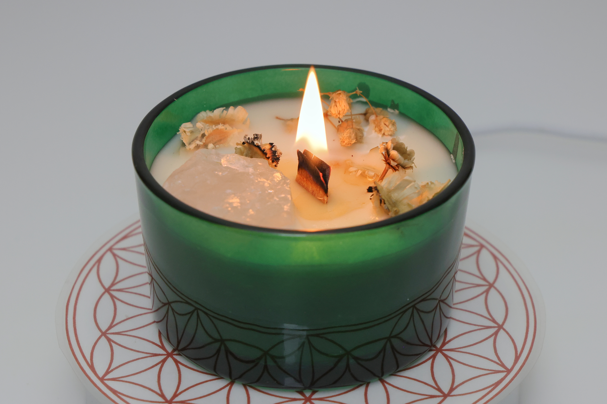 Grüne Duftkerze mit Holzdocht, dekorativen Blüten und einem eingebetteten Kristall, brennend auf einer weißen Unterlage mit rotem geometrischem Muster.
