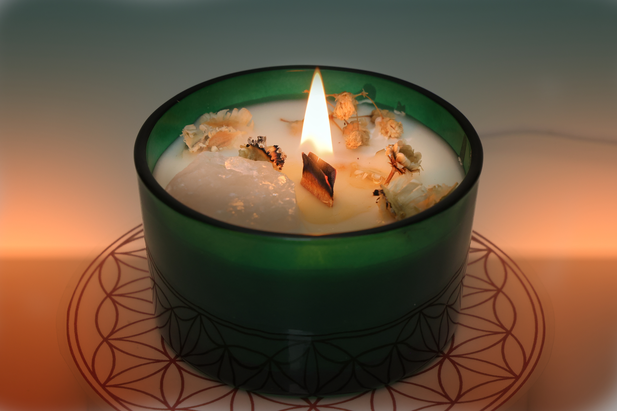 Grüne Duftkerze mit Holzdocht, dekorativen Blüten und einem eingebetteten Kristall, brennend auf einer weißen Unterlage mit rotem geometrischem Muster.