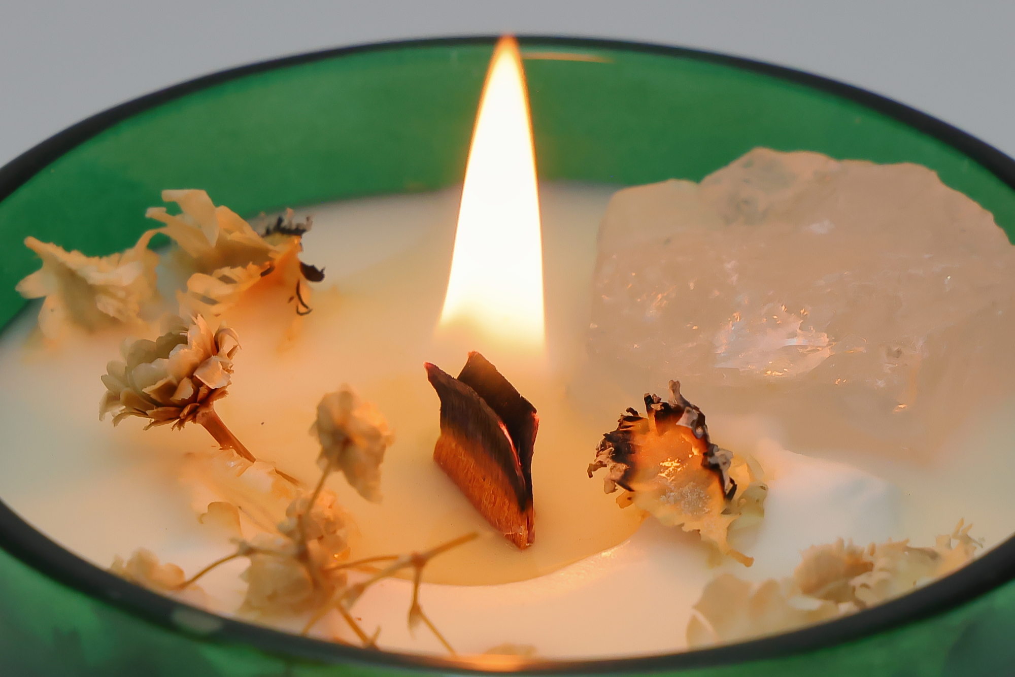 Grüne Duftkerze mit Holzdocht, dekorativen Blüten und einem eingebetteten Kristall, brennend auf einer weißen Unterlage mit rotem geometrischem Muster.