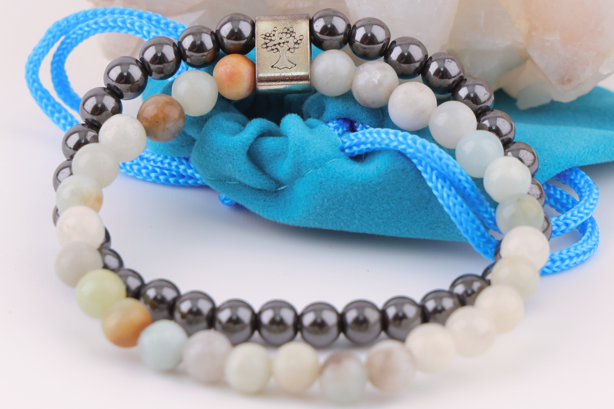 Sodalith‑Magnetarmband mit schwarzen und hellen Edelsteinperlen, dekorativ präsentiert auf einem klaren Bergkristall‑Cluster; davor ein türkisfarbenes Stoffbeutelchen mit blauen Zugbändern.
