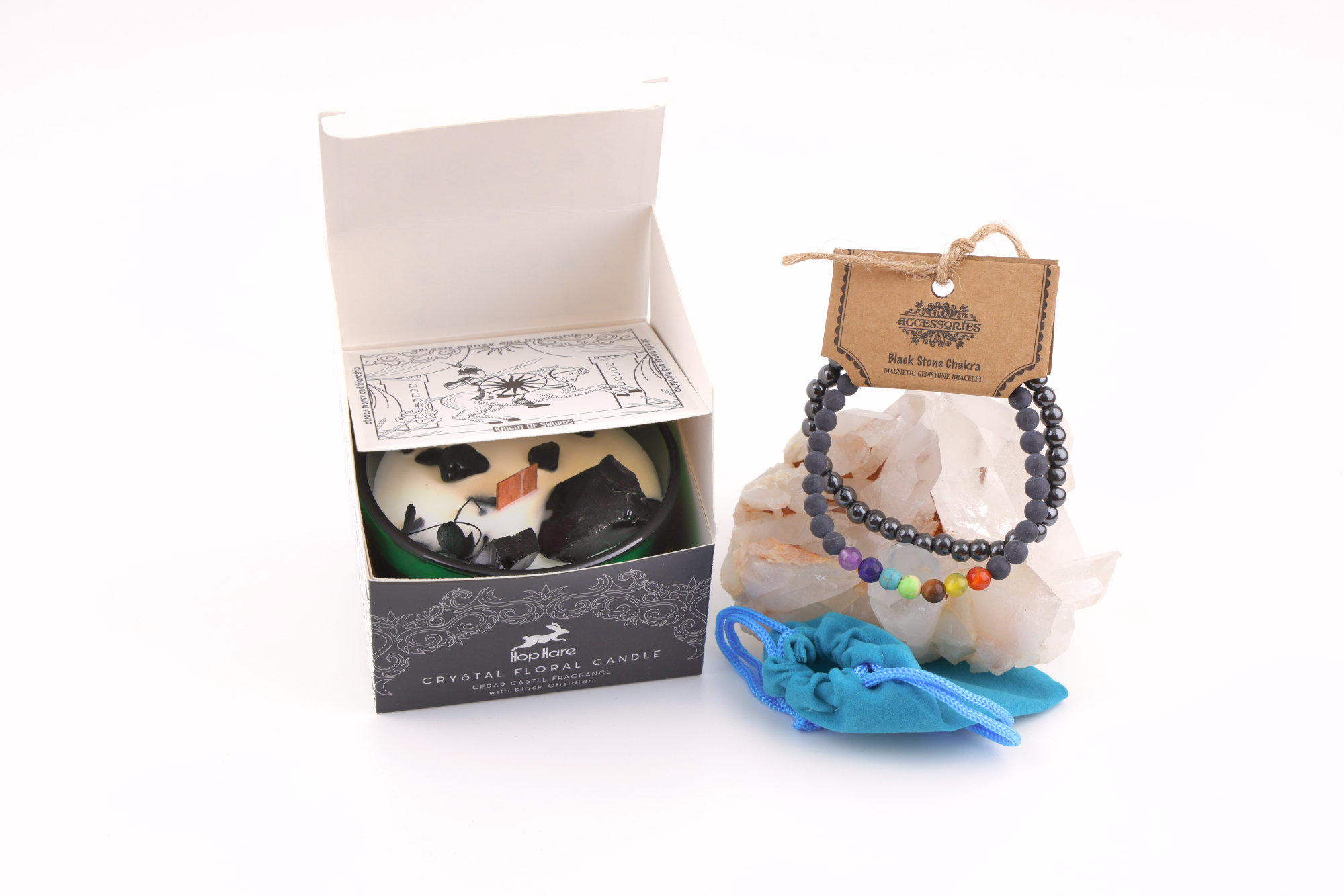Ein grünes Glas mit einer weißen Hop Hare „Der König der Schwerter“-Blumenkerze, dekoriert mit schwarzem Obsidian, Blättern und Holzdocht, daneben ein Schwarzstein‑Chakra‑Magnetarmband auf klaren Bergkristallen sowie ein türkisfarbener Stoffbeutel – alles auf weißem Hintergrund arrangiert.