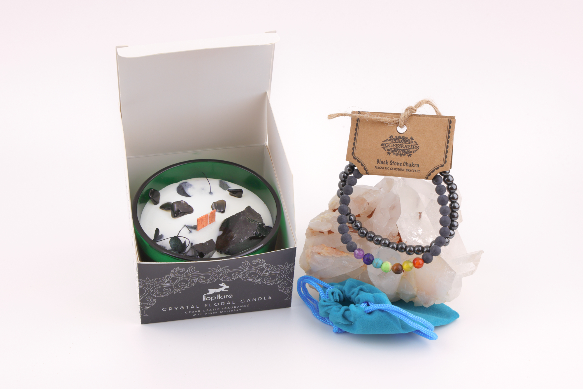 Ein grünes Glas mit einer weißen Hop Hare „Der König der Schwerter“-Blumenkerze, dekoriert mit schwarzem Obsidian, Blättern und Holzdocht, daneben ein Schwarzstein‑Chakra‑Magnetarmband auf klaren Bergkristallen sowie ein türkisfarbener Stoffbeutel – alles auf weißem Hintergrund arrangiert.
