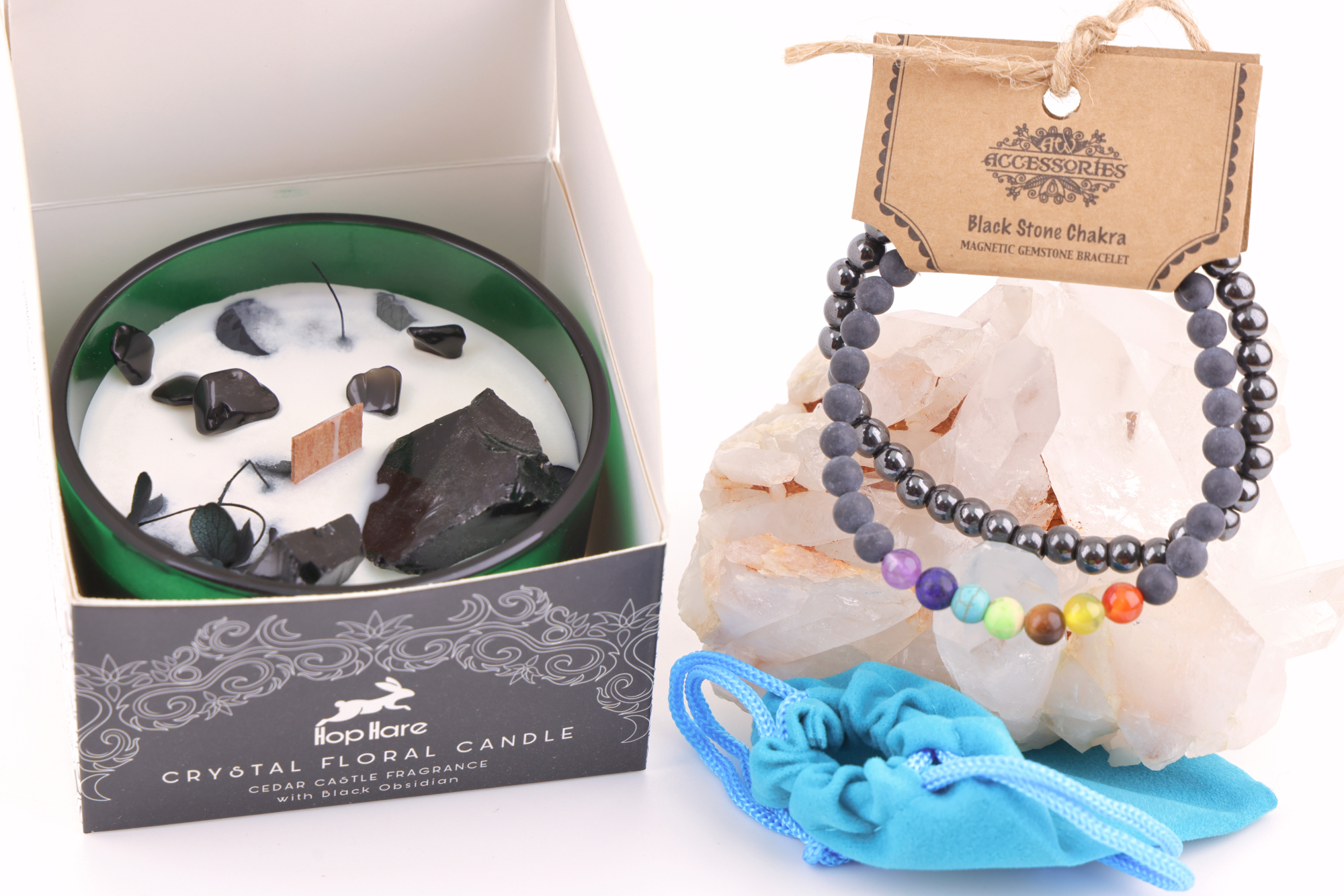 Ein grünes Glas mit einer weißen Hop Hare „Der König der Schwerter“-Blumenkerze, dekoriert mit schwarzem Obsidian, Blättern und Holzdocht, daneben ein Schwarzstein‑Chakra‑Magnetarmband auf klaren Bergkristallen sowie ein türkisfarbener Stoffbeutel – alles auf weißem Hintergrund arrangiert.