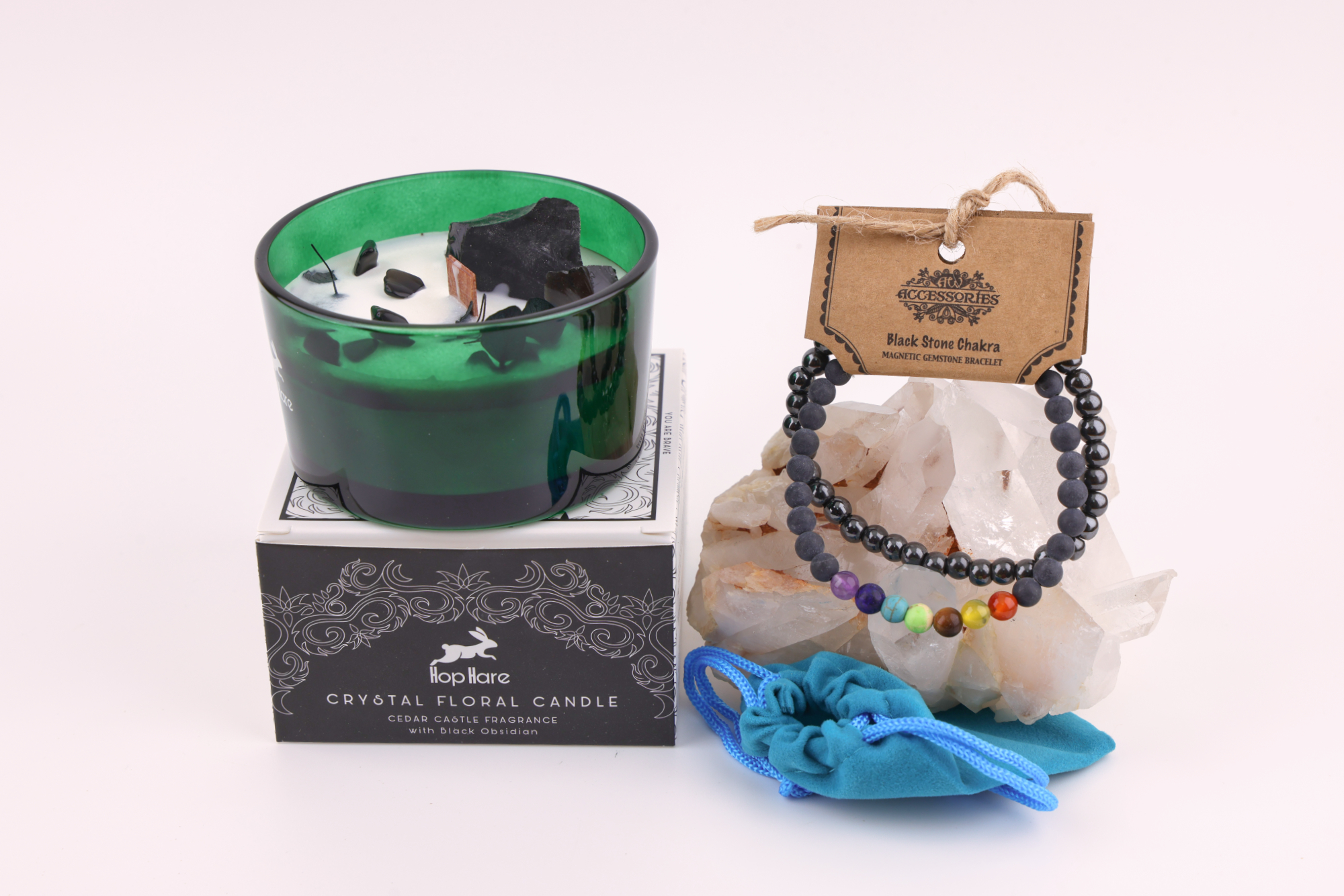 Ein grünes Glas mit einer weißen Hop Hare „Der König der Schwerter“-Blumenkerze, dekoriert mit schwarzem Obsidian, Blättern und Holzdocht, daneben ein Schwarzstein‑Chakra‑Magnetarmband auf klaren Bergkristallen sowie ein türkisfarbener Stoffbeutel – alles auf weißem Hintergrund arrangiert.