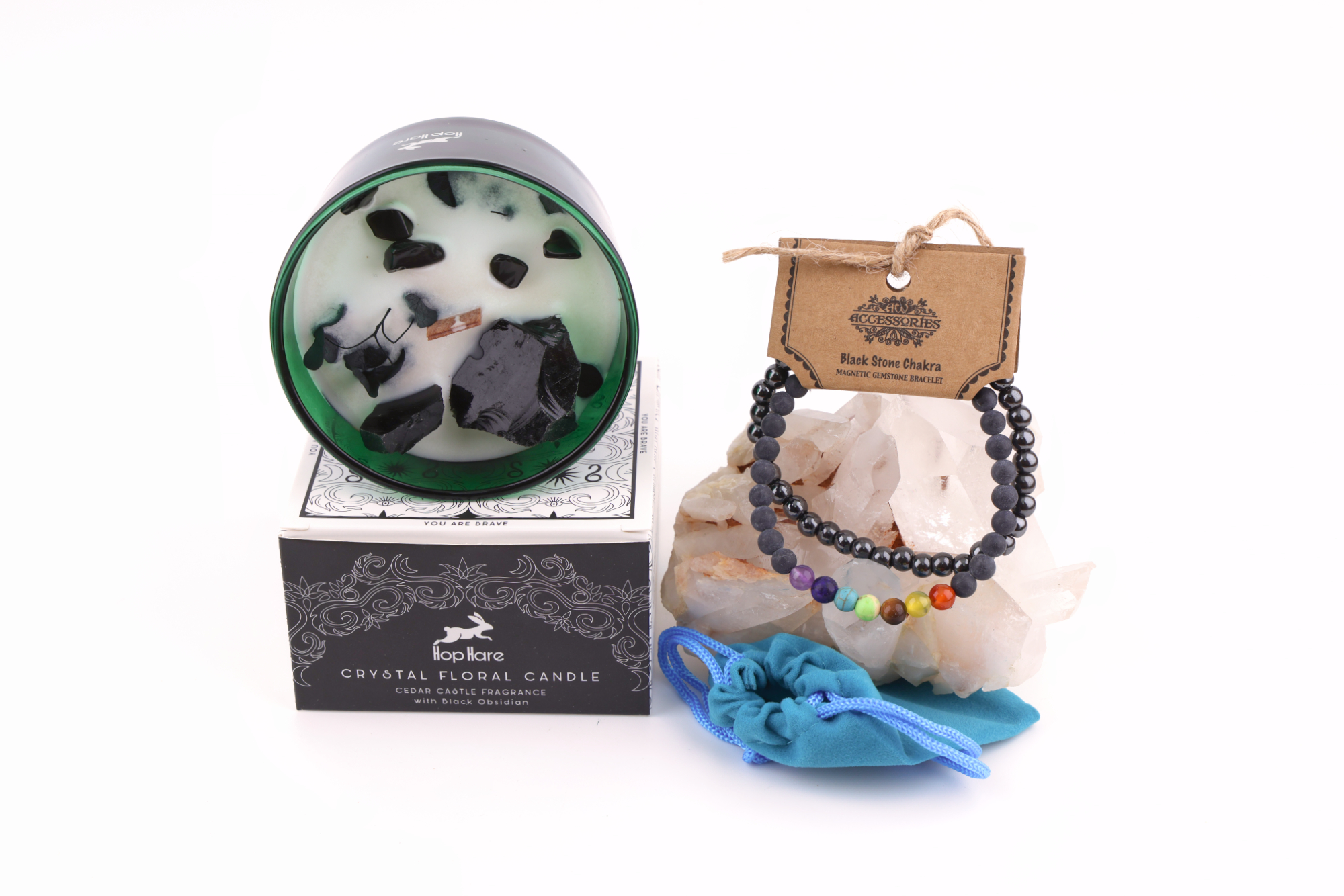 Ein grünes Glas mit einer weißen Hop Hare „Der König der Schwerter“-Blumenkerze, dekoriert mit schwarzem Obsidian, Blättern und Holzdocht, daneben ein Schwarzstein‑Chakra‑Magnetarmband auf klaren Bergkristallen sowie ein türkisfarbener Stoffbeutel – alles auf weißem Hintergrund arrangiert.