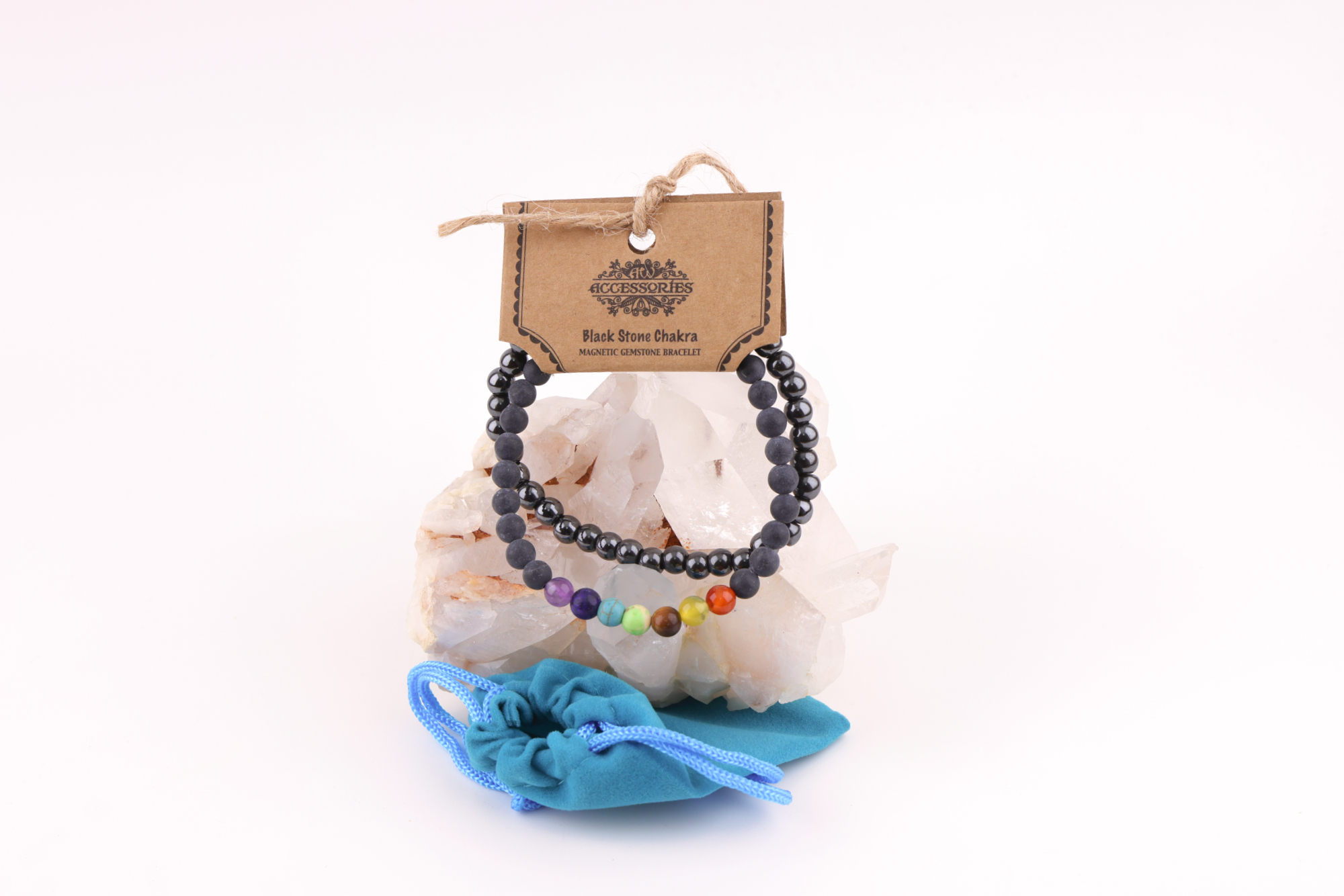 Ein Schwarzstein‑Chakra‑Magnetarmband aus schwarzen Perlen mit sieben farbigen Chakra‑Steinen, präsentiert auf einer klaren Bergkristall‑Druse; davor liegt ein kleiner türkisfarbener Stoffbeutel.
