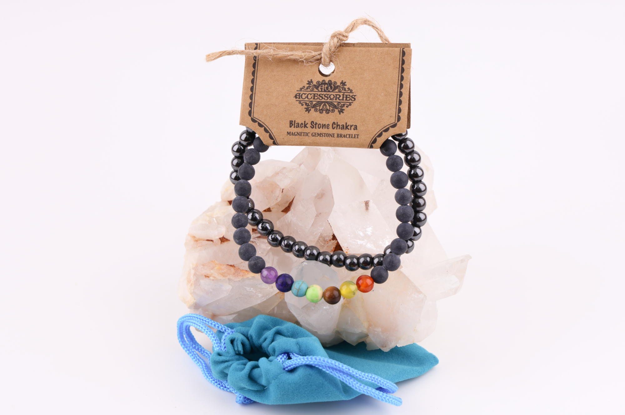 Ein Schwarzstein‑Chakra‑Magnetarmband aus schwarzen Perlen mit sieben farbigen Chakra‑Steinen, präsentiert auf einer klaren Bergkristall‑Druse; davor liegt ein kleiner türkisfarbener Stoffbeutel.
