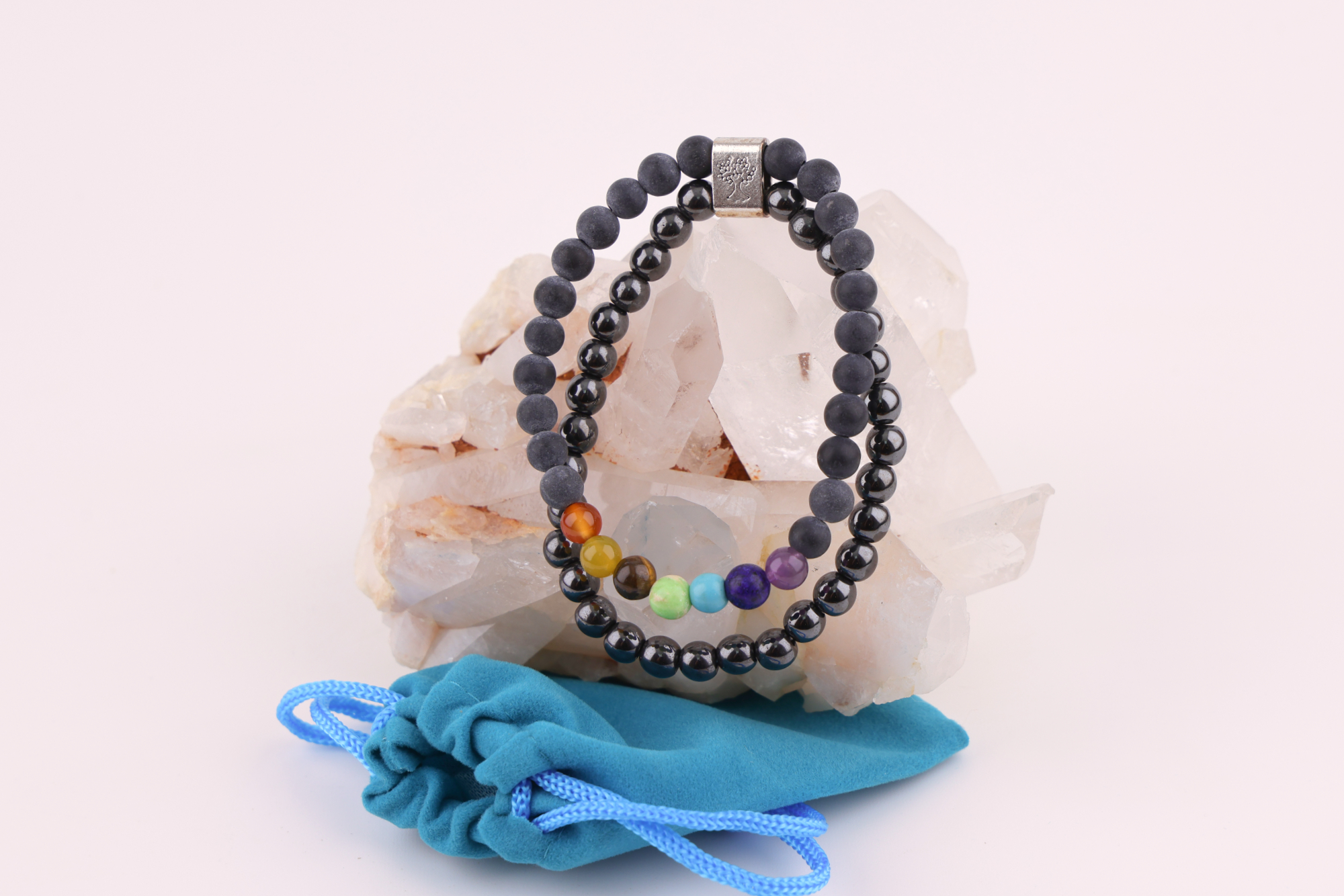 Ein Schwarzstein‑Chakra‑Magnetarmband aus schwarzen Perlen mit sieben farbigen Chakra‑Steinen, präsentiert auf einer klaren Bergkristall‑Druse; davor liegt ein kleiner türkisfarbener Stoffbeutel.