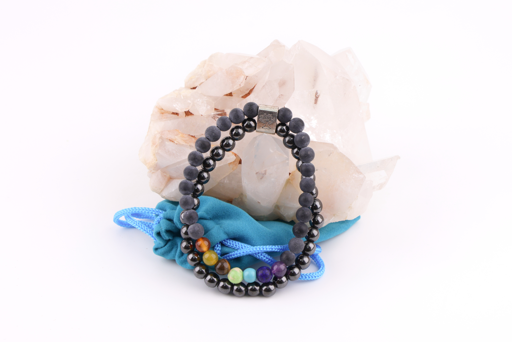 Ein Schwarzstein‑Chakra‑Magnetarmband aus schwarzen Perlen mit sieben farbigen Chakra‑Steinen, präsentiert auf einer klaren Bergkristall‑Druse; davor liegt ein kleiner türkisfarbener Stoffbeutel.