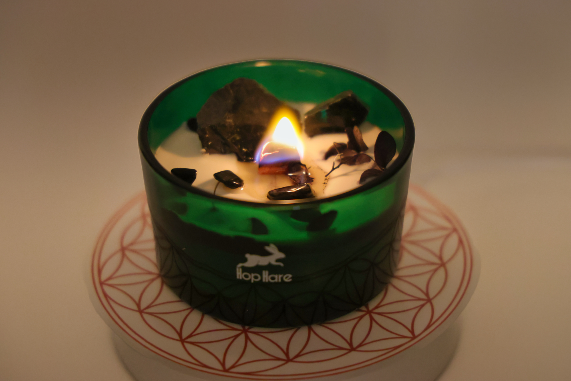   
Eine brennende Hop Hare Kerze in einem grünen Glas, mit weißem Wachs, dunklen Steinen und kleinen Pflanzenstücken, platziert auf einer runden Unterlage mit rot geometrischem Blütenmuster.
