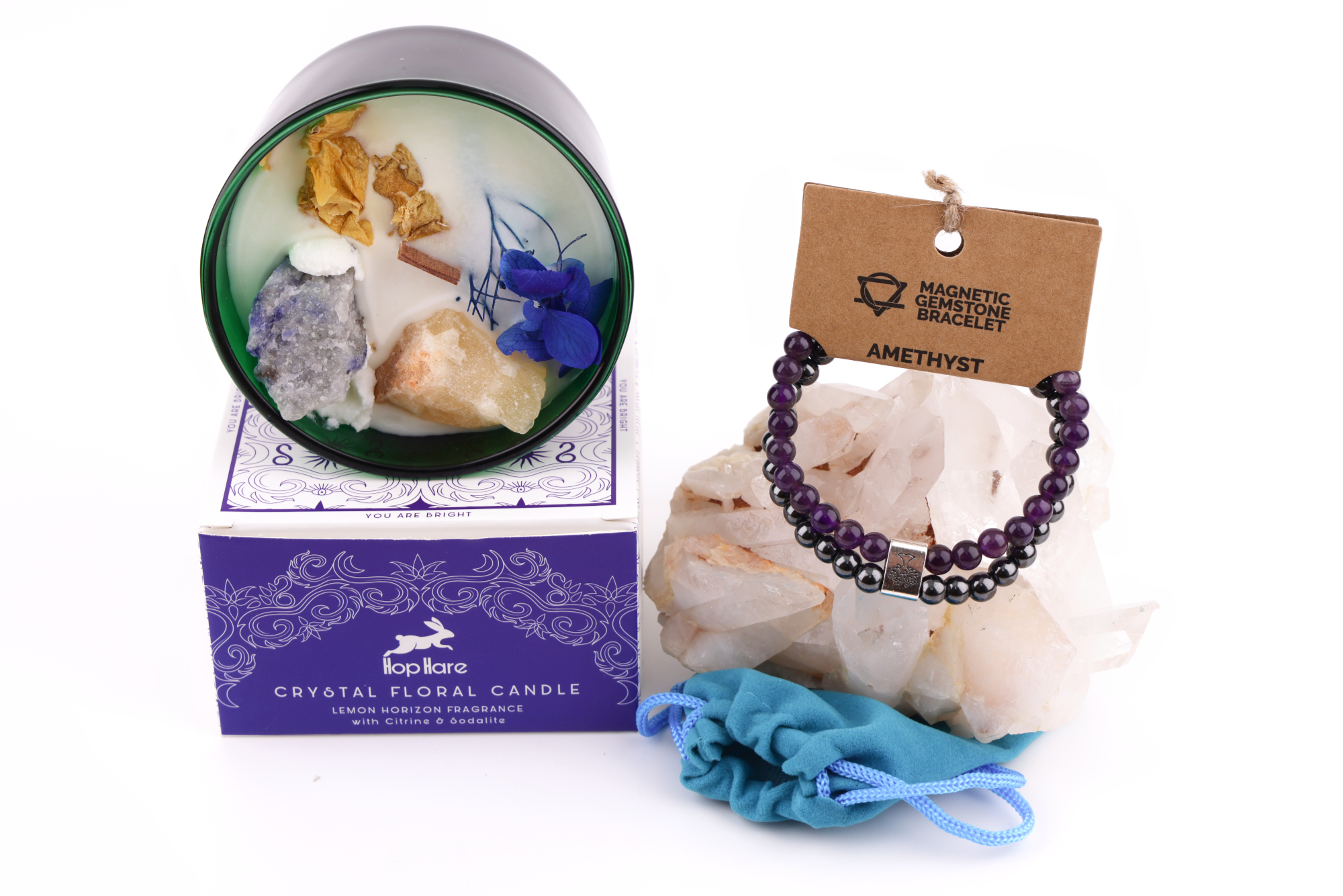 Grüne Hop‑Hare Duftkerze mit Citrin‑ und Amethyst‑Steinen sowie Blüten, daneben die lilafarbene Produktbox; rechts ein Amethyst‑Magnetarmband auf brauner Karte, präsentiert auf Bergkristallen mit türkisfarbenem Beutel.
