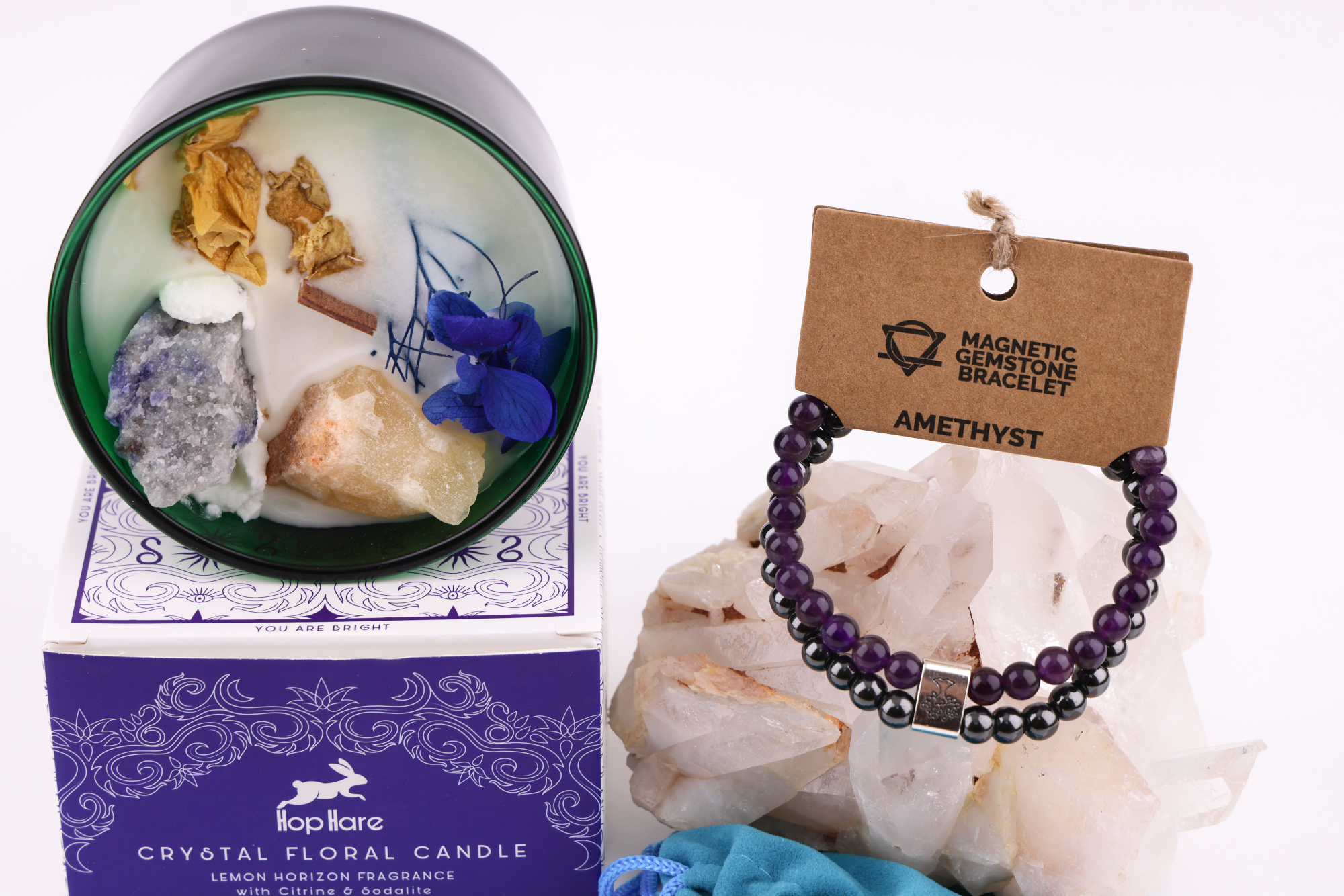 Grüne Hop‑Hare Duftkerze mit Citrin‑ und Amethyst‑Steinen sowie Blüten, daneben die lilafarbene Produktbox; rechts ein Amethyst‑Magnetarmband auf brauner Karte, präsentiert auf Bergkristallen mit türkisfarbenem Beutel.
