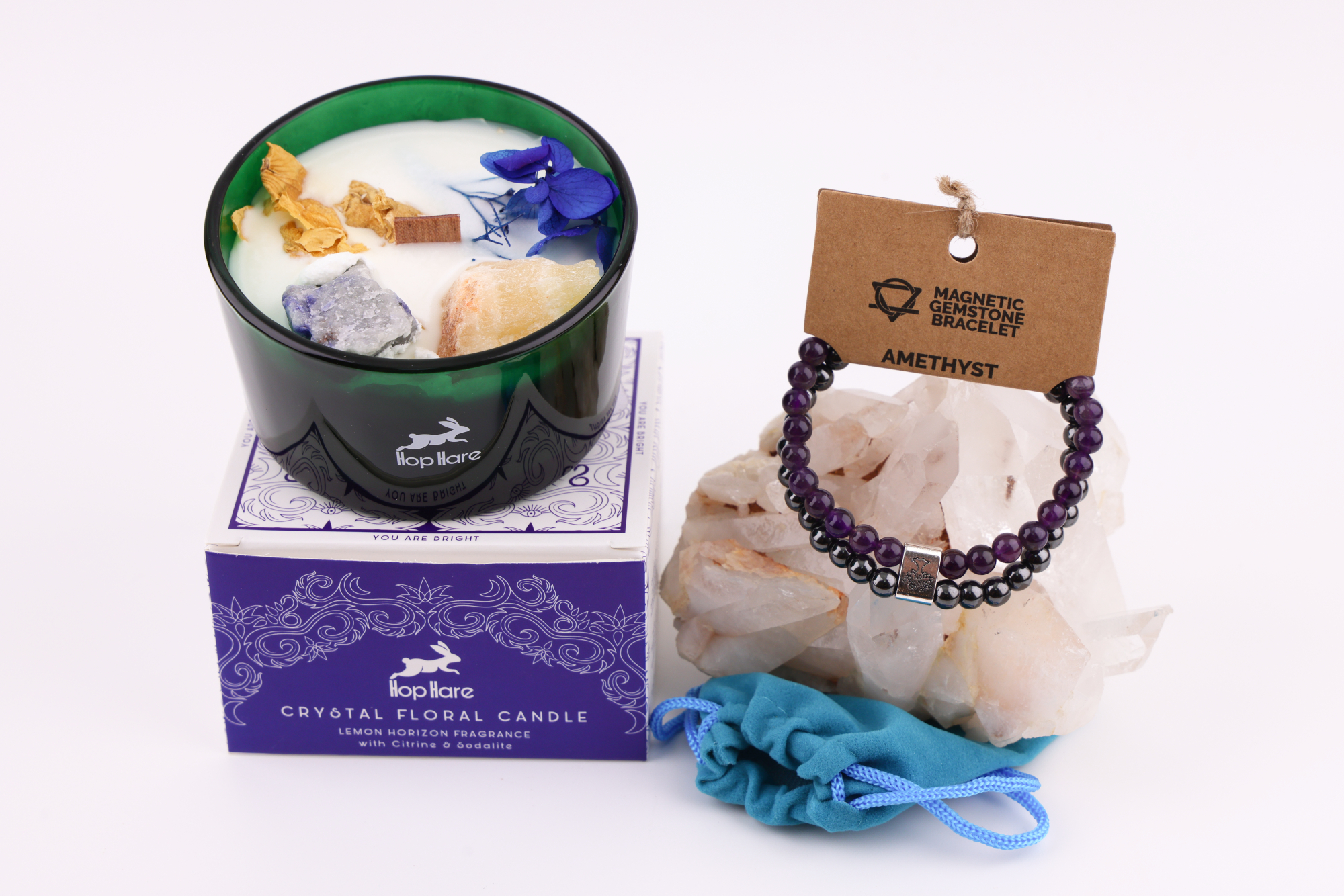 Grüne Hop‑Hare Duftkerze mit Citrin‑ und Amethyst‑Steinen sowie Blüten, daneben die lilafarbene Produktbox; rechts ein Amethyst‑Magnetarmband auf brauner Karte, präsentiert auf Bergkristallen mit türkisfarbenem Beutel.