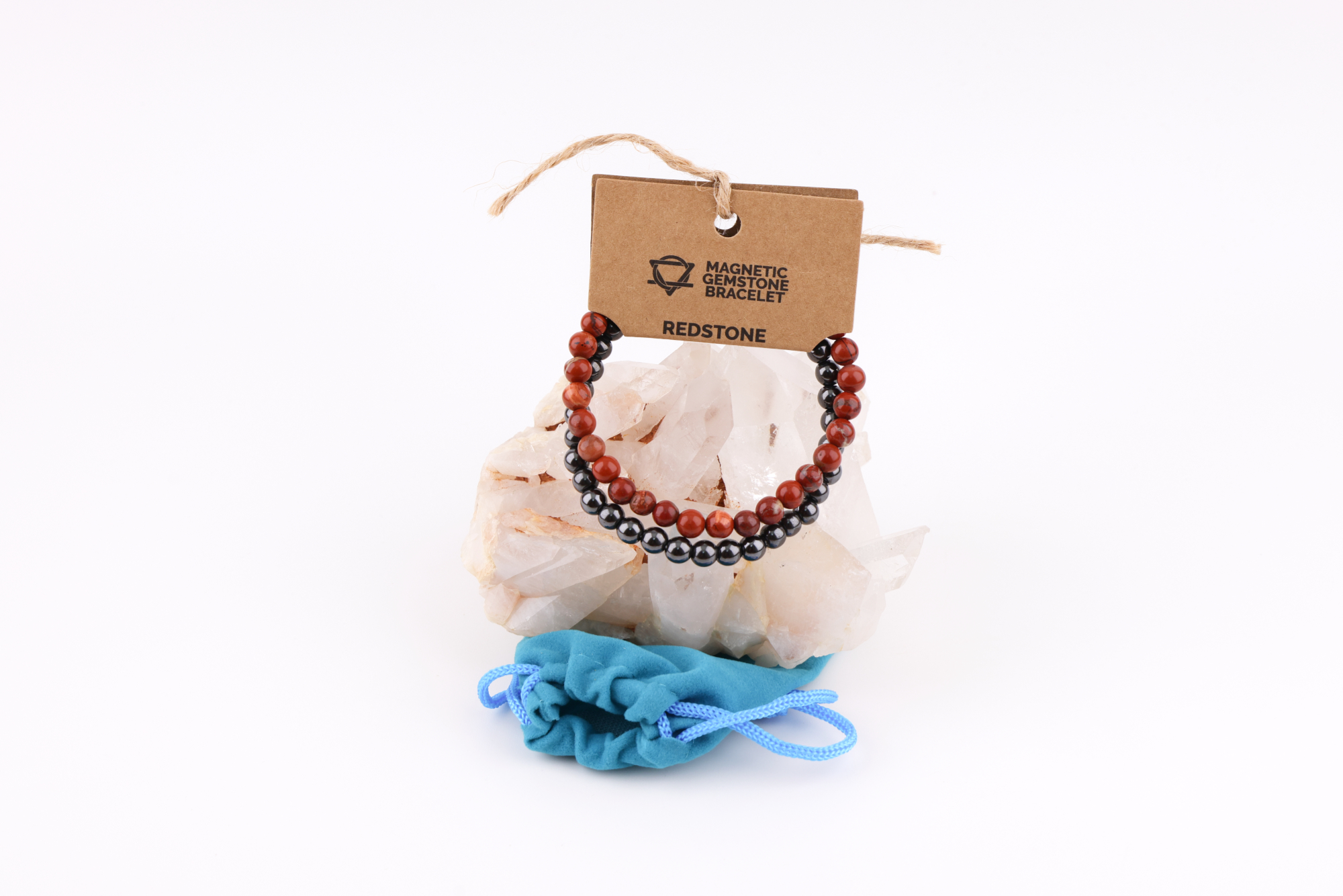 Ein Redstone‑Magnetarmband aus roten und dunklen Metallperlen, präsentiert auf einer klaren Bergkristall‑Druse; davor liegt ein kleiner blauer Stoffbeutel.