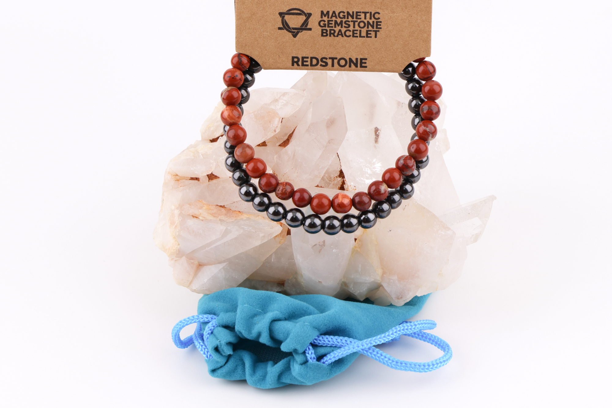 Ein Redstone‑Magnetarmband aus roten und dunklen Metallperlen, präsentiert auf einer klaren Bergkristall‑Druse; davor liegt ein kleiner blauer Stoffbeutel.