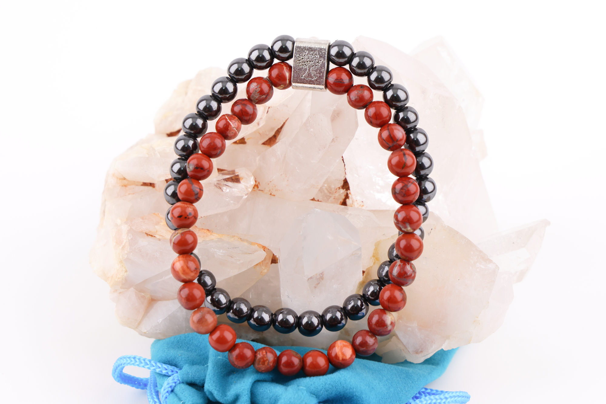 Ein Redstone‑Magnetarmband aus roten und dunklen Metallperlen, präsentiert auf einer klaren Bergkristall‑Druse; davor liegt ein kleiner blauer Stoffbeutel.
