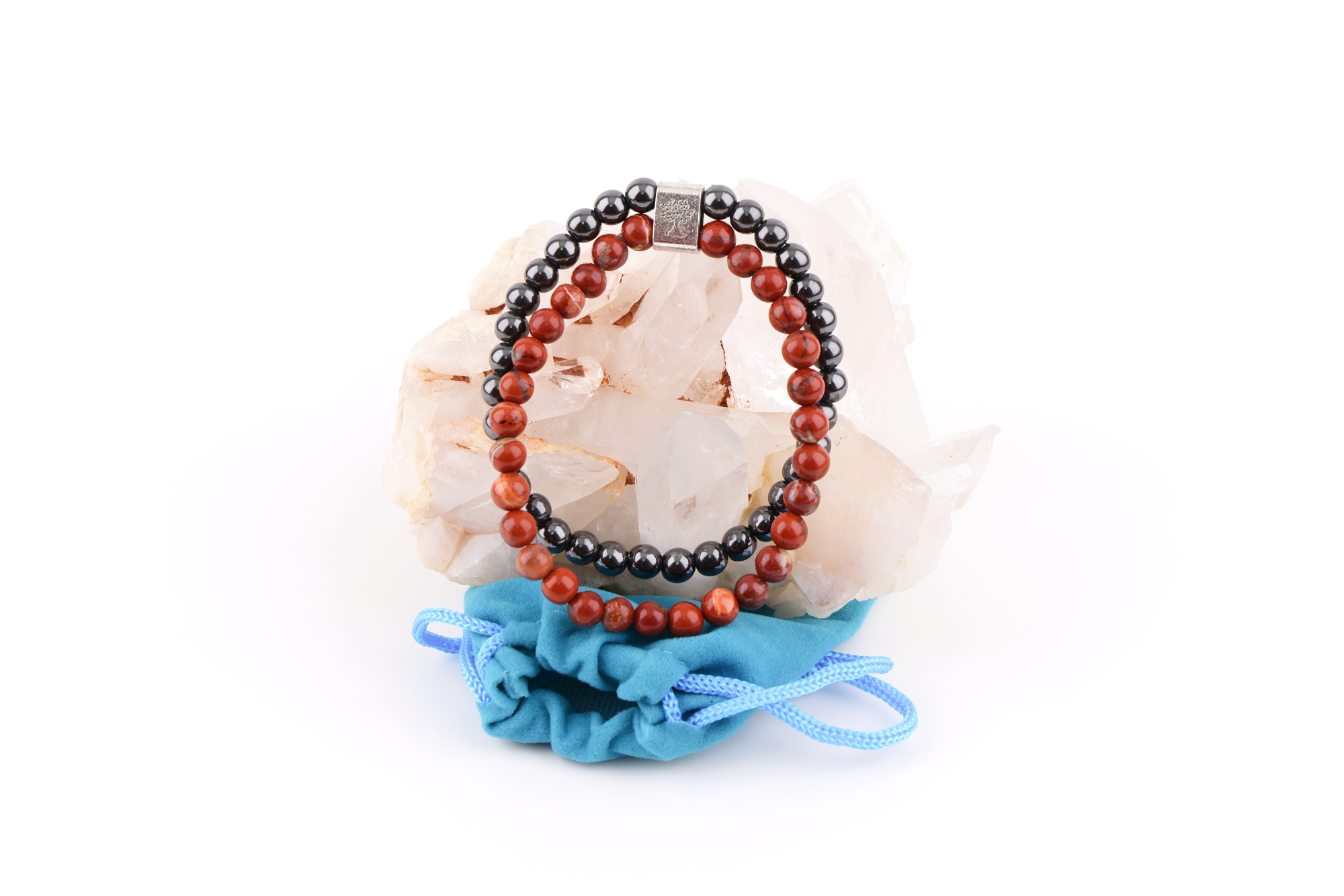 Ein Redstone‑Magnetarmband aus roten und dunklen Metallperlen, präsentiert auf einer klaren Bergkristall‑Druse; davor liegt ein kleiner blauer Stoffbeutel.