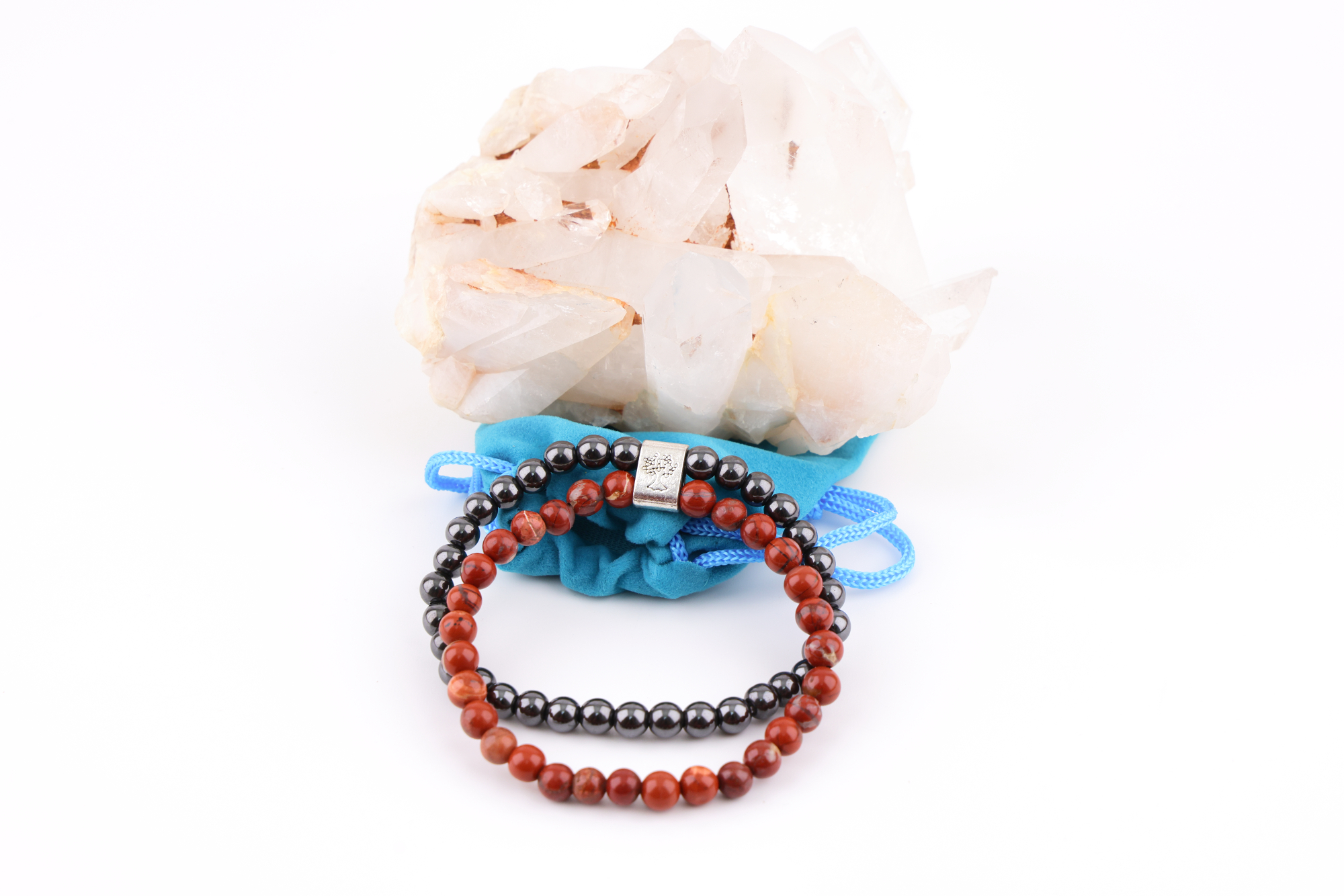 Ein Redstone‑Magnetarmband aus roten und dunklen Metallperlen, präsentiert auf einer klaren Bergkristall‑Druse; davor liegt ein kleiner blauer Stoffbeutel.