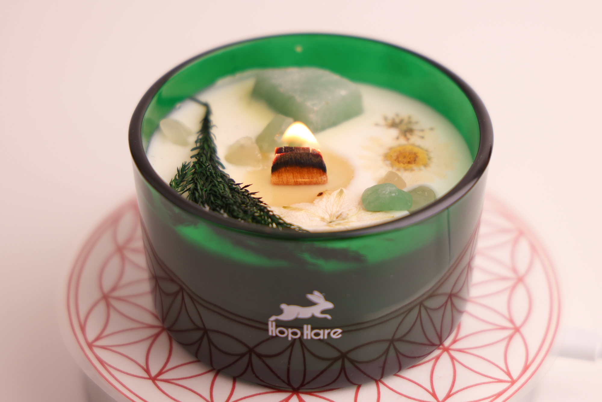„Brennende Hop‑Hare‑Kerze mit Blüten und grünen Steinen im Glas auf roter Musterplatte.