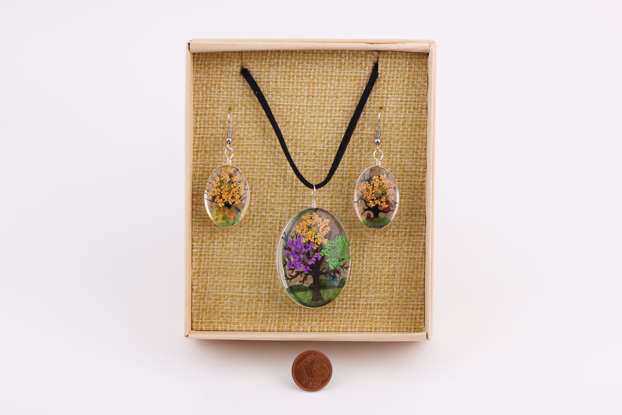 Baum‑des‑Lebens Schmuckset mit blauem Baum‑Motiv, bestehend aus Halsanhänger und Ohrringen in beige Geschenkbox, daneben eine Münze als Größenvergleich.