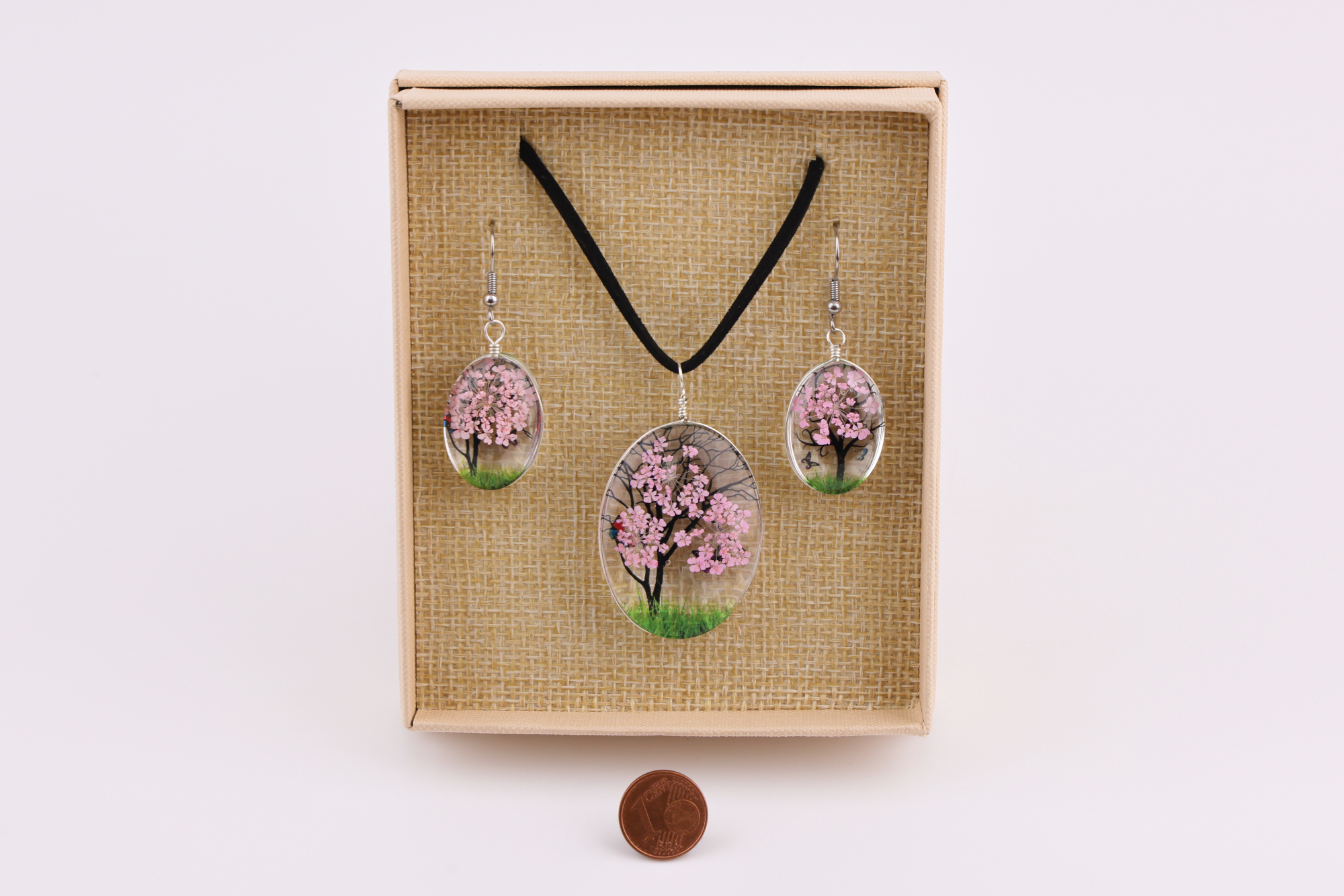 Baum‑des‑Lebens Schmuckset mit rosa Baum‑Motiv, bestehend aus Halsanhänger und Ohrringen in beige Geschenkbox, daneben eine Münze als Größenvergleich.