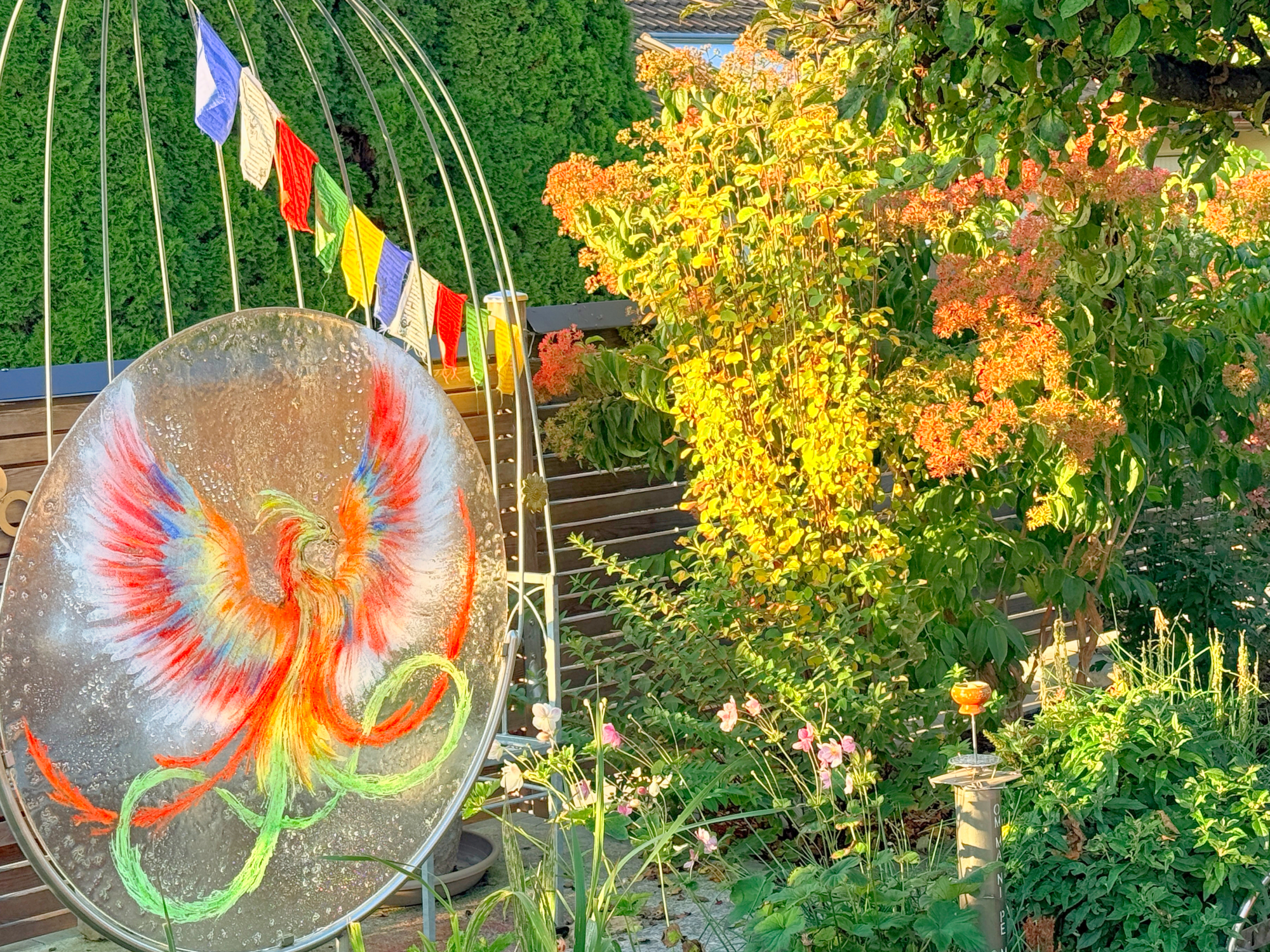 Tibetische Gebetsfähnchen in fünf Farben hängen diagonal im Garten vor grünen Sträuchern